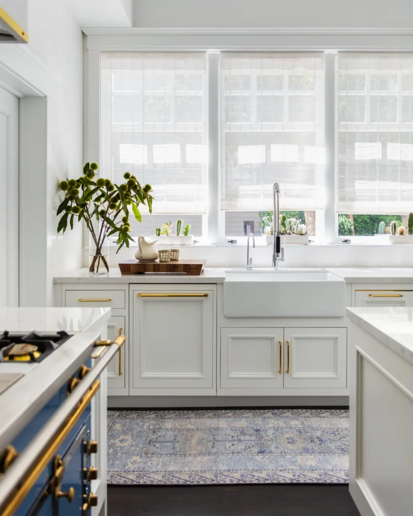 Elegant white kitchen with gold hardware, farmhouse sink, and sunlit windows with soft shades