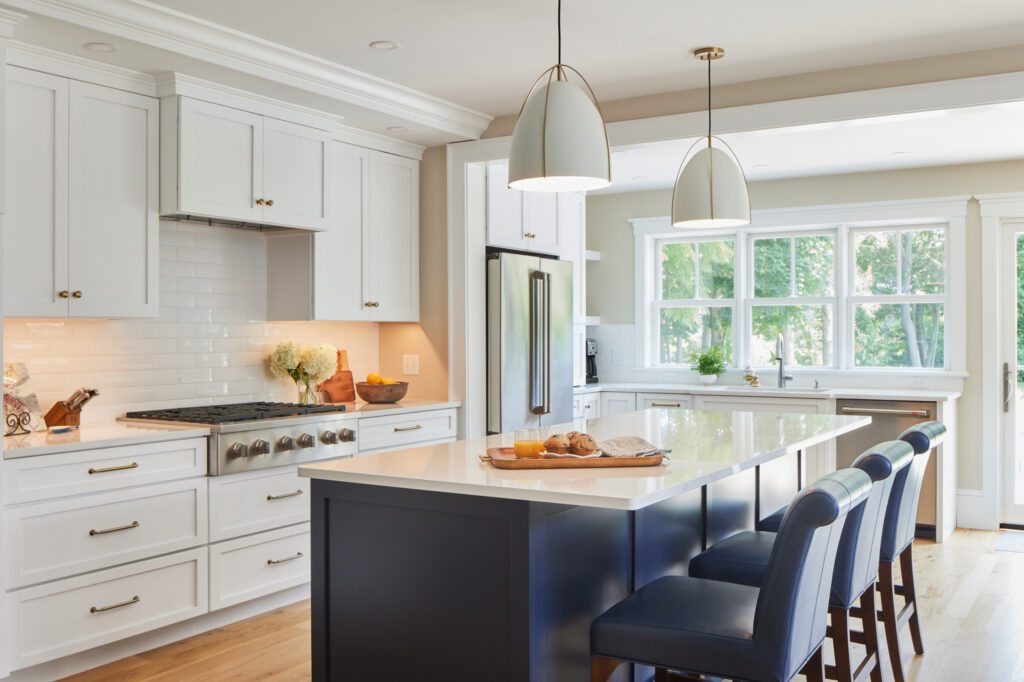 Modern kitchen with white shaker cabinets, navy blue island, pendant lighting, stainless steel appliances, and a large window overlooking greenery