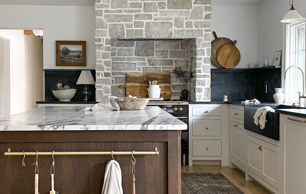 Cozy rustic kitchen with a natural stone hood surround, marble-topped island, white shaker cabinets, and black countertops.