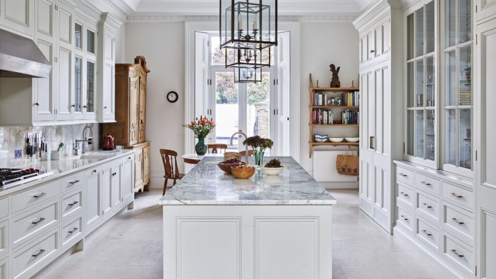 A bright, classic-style kitchen featuring marble countertops, tall white cabinetry, and a central island with flowers and fruit bowls.