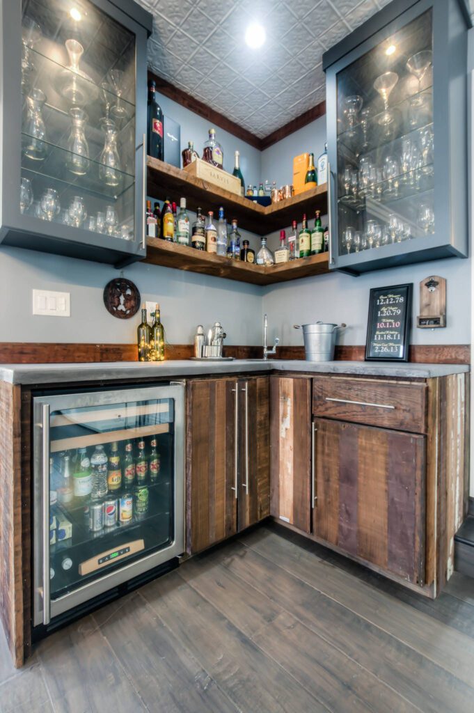 A stylish corner home bar featuring rustic wood cabinetry, floating wood shelves stocked with liquor bottles, glass-front upper cabinets with glassware, and a built-in beverage cooler.