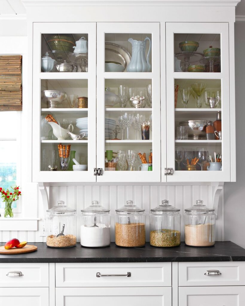 White kitchen cabinet with glass doors showcasing dishes and glassware, and five large glass storage jars filled with pantry items on a black countertop