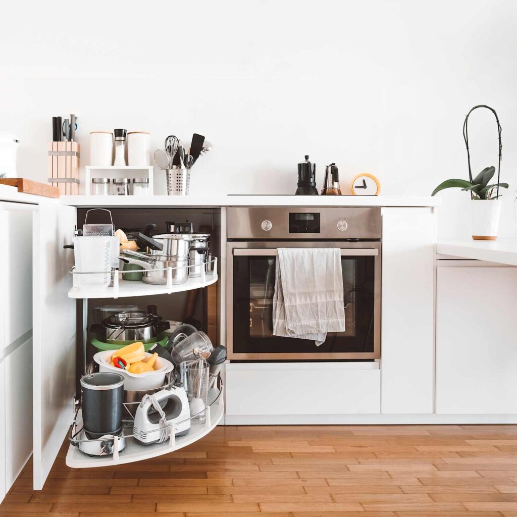 White modern kitchen with a corner pull-out cabinet fully extended, holding cookware, utensils, and kitchen gadgets.