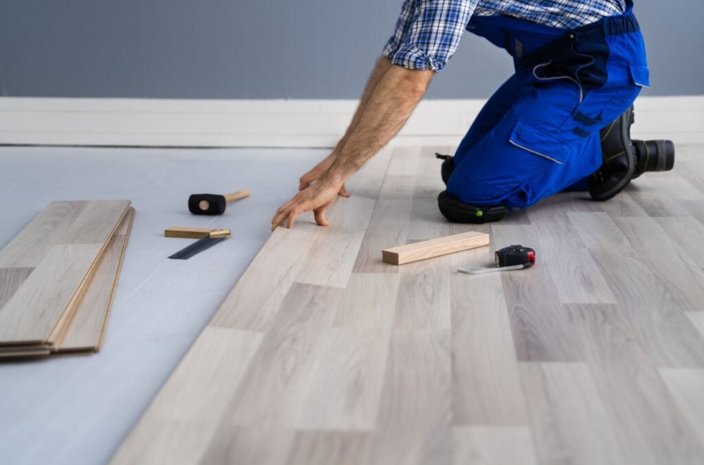 A flooring contractor kneeling and installing light-colored vinyl plank flooring using tools.