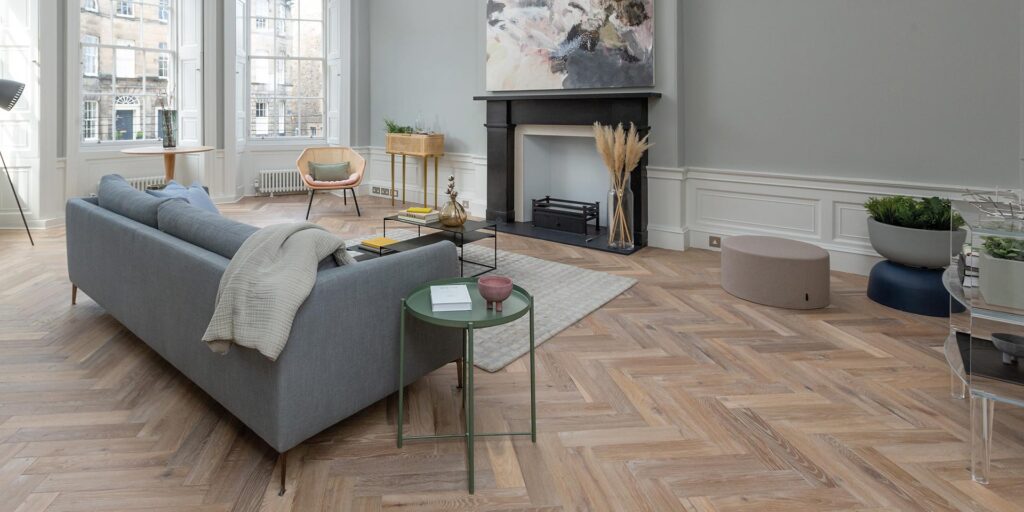 A spacious, light-filled living room featuring herringbone-patterned wood flooring and modern minimalist decor.