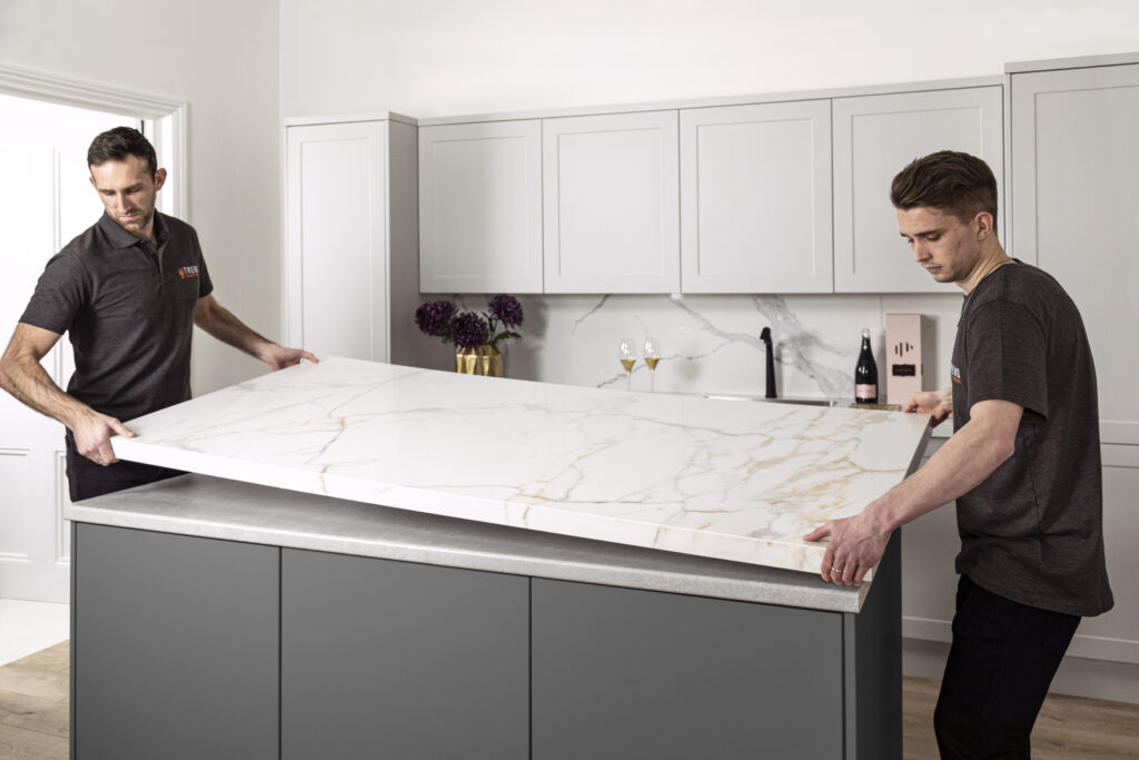 Two workers installing a white marble-look countertop on a gray kitchen island