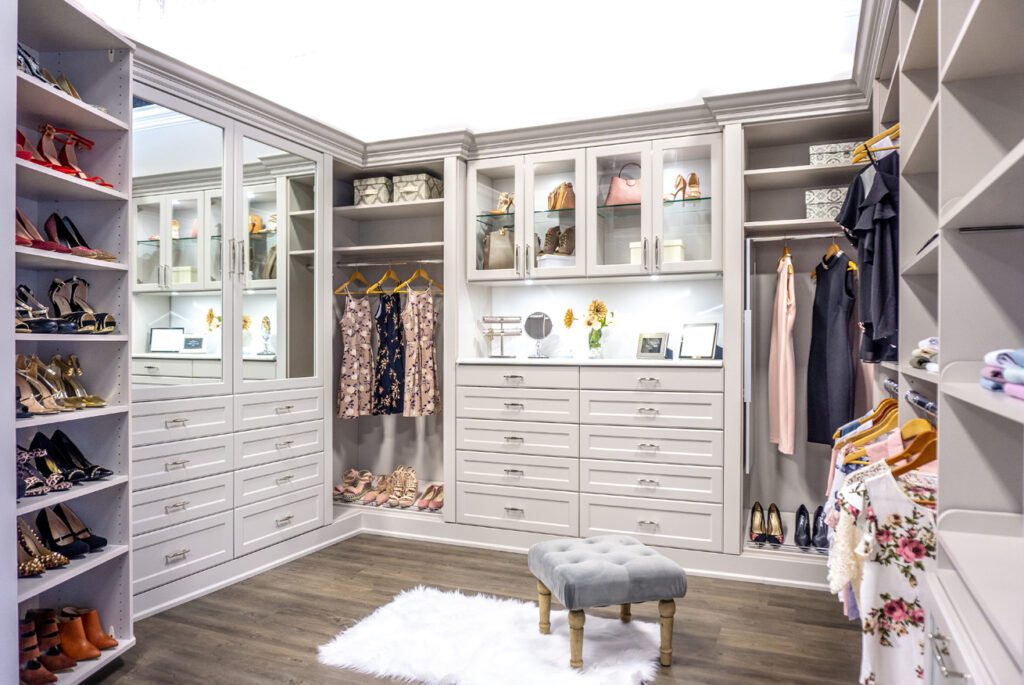 Spacious walk-in closet with white cabinetry, glass display cabinets, and a soft gray ottoman