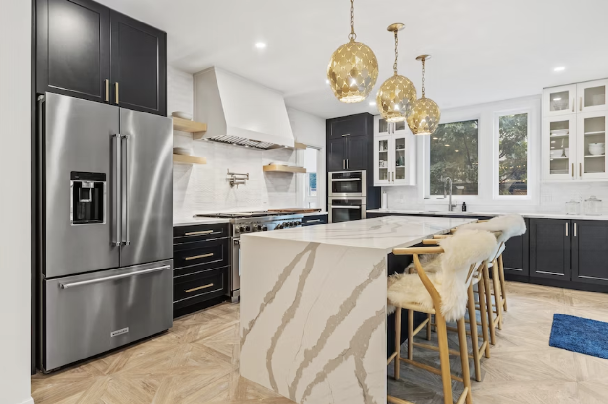 Modern kitchen with black cabinets, Carrara marble waterfall island, gold pendant lights, and wood flooring.