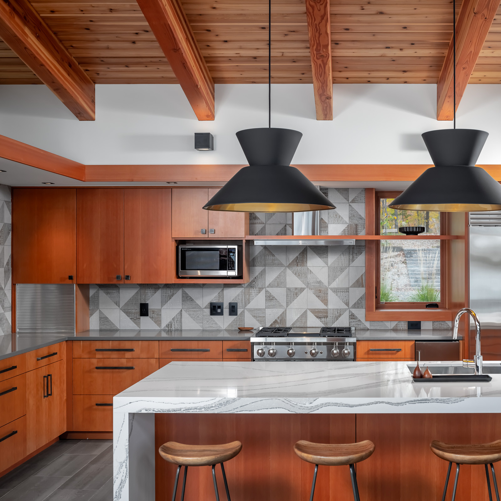 Modern kitchen with wood cabinets, marble island, geometric backsplash, and black pendant lights under a wood-beamed ceiling.