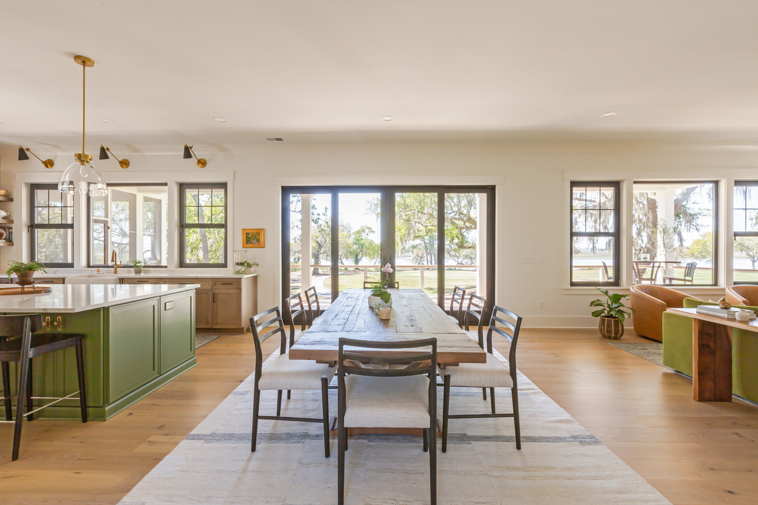Open kitchen and dining area with green island, wooden dining table, and large windows offering outdoor views.