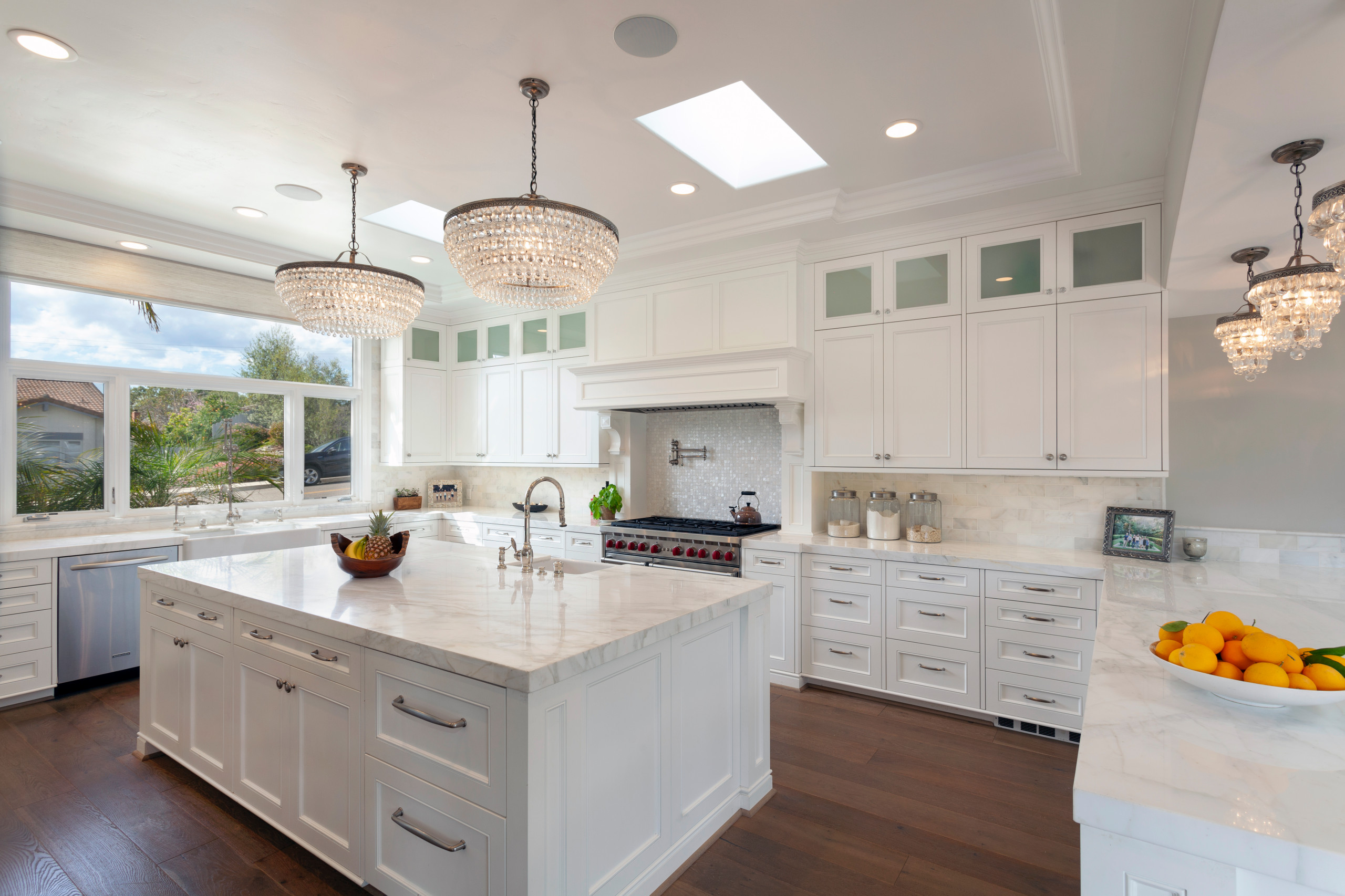 Luxurious white kitchen with marble countertops, crystal chandeliers, and stainless-steel appliances.