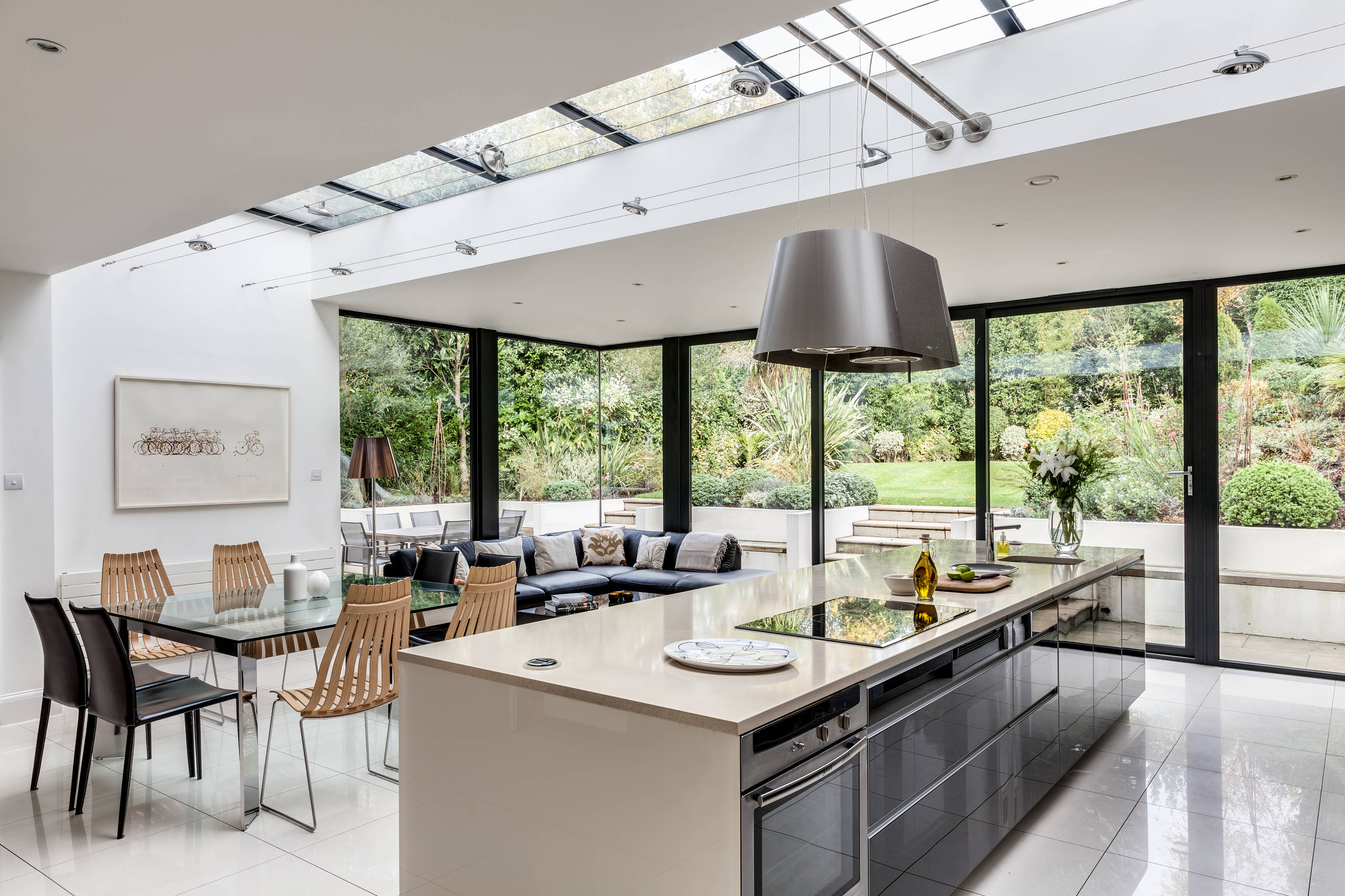 Modern open-plan kitchen with large glass windows, sleek island, and dining area overlooking a garden.