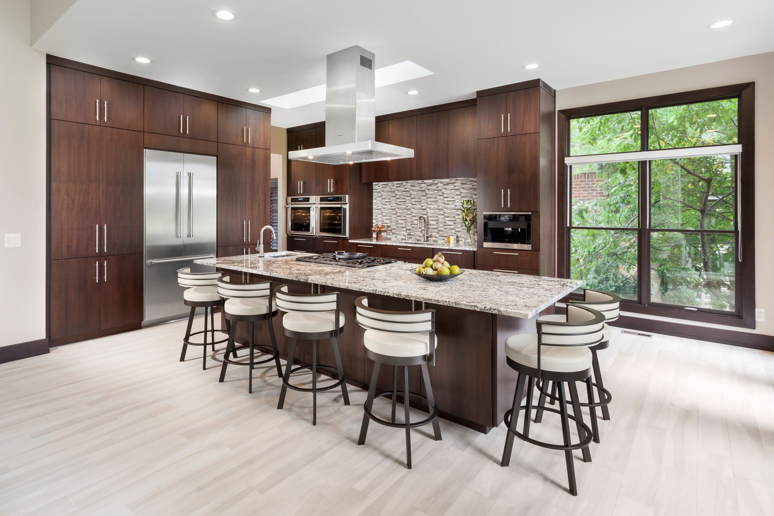 Modern kitchen with dark wood cabinetry, a large granite island, and stainless-steel appliances under bright recessed lighting.