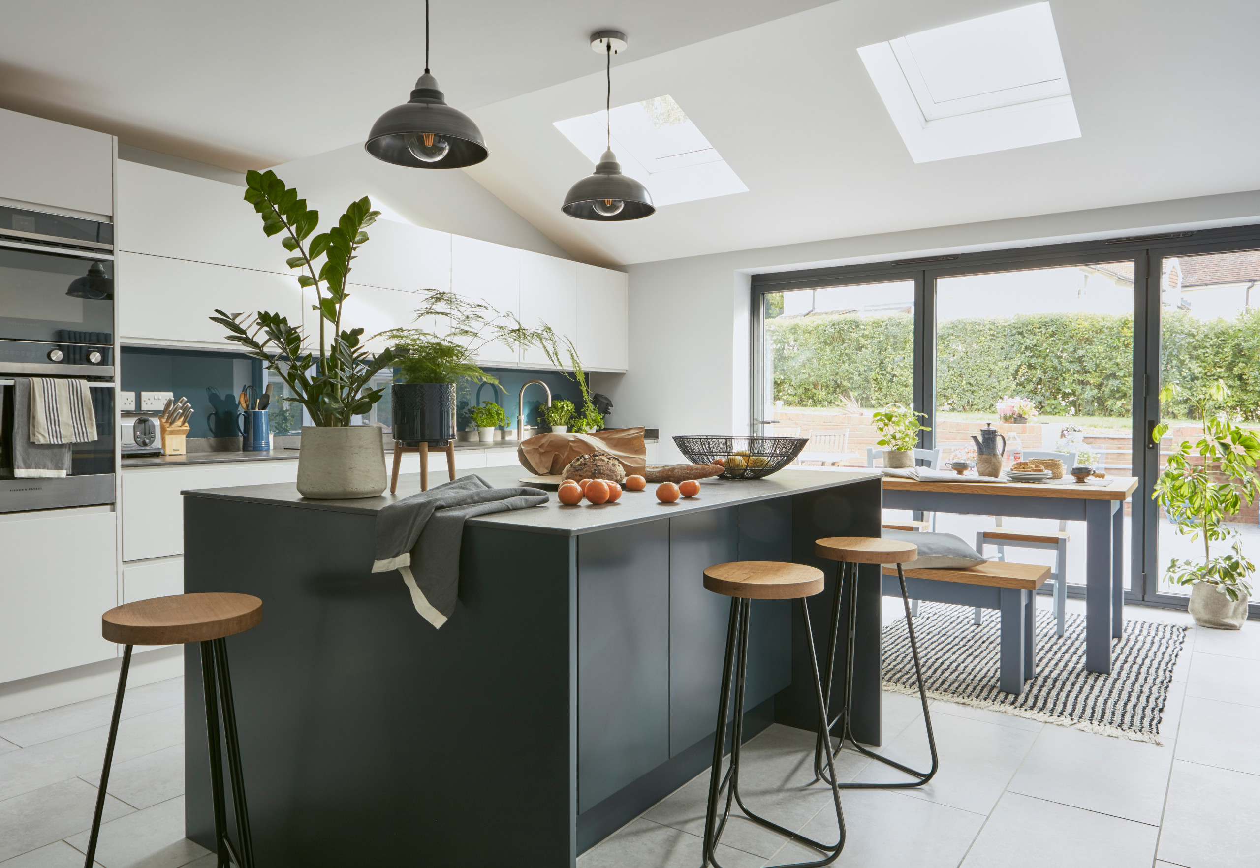 Modern kitchen interior featuring a stylish island with wooden stools, pendant lights, and lush green plants under skylights.