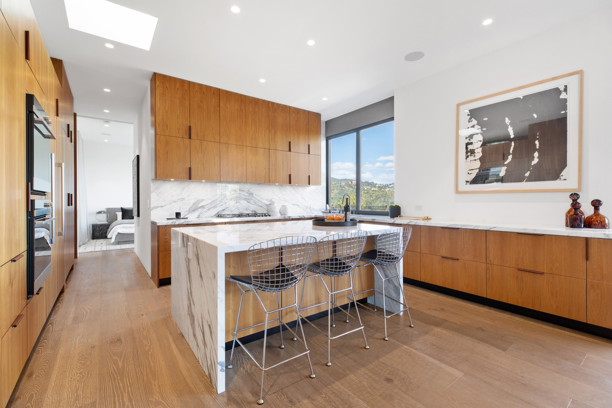 Modern kitchen with natural wood cabinetry, marble island, and sleek metal bar stools under recessed lighting.